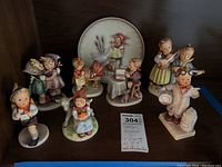 Eight Goebel Hummel porcelain figurines and one commemorative plate displayed together on a shelf.