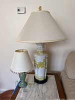 Full view of two table lamps sitting on a marble-topped table: a tall white and gold floral design lamp with a large beige shade and a small green ceramic lamp with a white fringed shade.