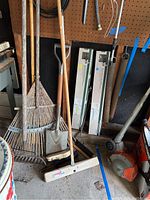 Various garden tools with wooden handles including rakes, shovels, and a broom leaning against a pegboard wall with boxes and other items present.