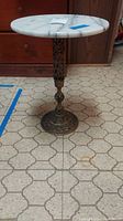 Full view of the round marble top brass base stand placed on patterned floor in front of wooden furniture. Shows entire stand with visible wear and surface detail.