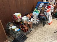 Black metal chaise lounge on wheels, shown with various gardening pots and decorations on top (not included). Visible against wood-paneled wall and hexagonal floor tiles.