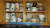 Cabinet shelf with 15 ceramic and pottery mugs in assorted designs, some with logos and floral patterns.