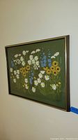 Full view of first framed needlework floral artwork with metal frame showing a variety of embroidered daisies on green fabric.