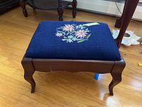 Side and top perspective view of the wood stool with needlepoint cushion, showing curved legs and general condition.