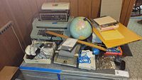 Photo shows the full lot of vintage office electronics including IBM electric typewriter, laminator, globe, cables, and various office supplies arranged on blue cart.