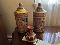 Photo of two lidded pottery canisters with painted designs and one small decorative vase on a wooden surface.