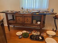 Dining room server with sliding top closed, two door compartments, open shelves both sides, and two bottom drawers. Some dishware and decorative items shown on and around it, not included.