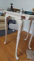 Side view of white painted wooden side table showing curved carved legs and ornamental detailing on corner and edges. Some wear marks are visible.
