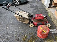 Photo of red Toro push mower with fabric grass collection bag and red metal gas can on asphalt surface.