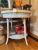 Full view of round wicker table showing woven wicker sides and legs, solid non-wicker top surface, and bottom wicker shelf with items placed on and under it.