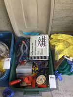 Overview photo showing a green plastic bin filled with assorted clamps including a boxed 12-piece spring clamp set, handheld clamps with red handles, purple plastic clamps, black clamps with orange tips in blue mesh bag, and other items.