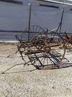 Overall side view showing large cast iron farm implement with two large spoked wheels and mechanical parts, under daylight next to a white tarp background.