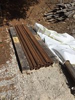 10 long heavy gauge steel pipes stacked on a wooden pallet outside, showing uniform rust and some dirt on surface.