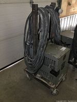 Side and top view of gray metal transformer unit on rusty metal cart with four caster wheels, thick coiled heavy-duty electrical cables draped over cart sides.