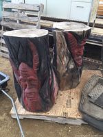 Photo of two large solid wood carvings with red horned faces carved into wood logs with black painted flames or hair design, double-sided faces visible.