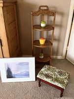 Oak 3-tiered shelf holding small containers, small floral upholstered footstool, framed mountain print leaning against wall