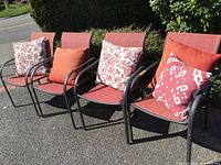 Four patio chairs arranged outside with brick red nylon seats and backs, black metal frames, and complementing pillows in various red and white patterns on them.