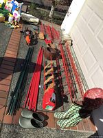 Full view of gardening tools, tomato vine stands, boots, and hedge trimmer laid out on ground next to garage.
