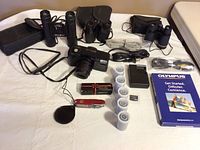 Lot contents showing Olympus camera, binoculars, film rolls, multi-tool knife and various cables and accessories.