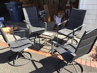 Four black swivel patio chairs arranged around small square glass-top table outside garage.