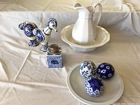 Full lot showing all items arranged on a cloth background: three blue and white spheres on a white plate, a blue and white ceramic horse figurine, white ceramic pitcher and basin, and a decorative box with metal lid.