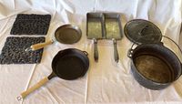 Photo showing all cookware items spread out including two cast iron frying pans with wooden handles, a cast iron Dutch oven with lid, two rectangular metal pans, and two trivet stands.