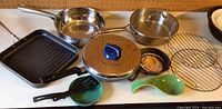 Photo showing stainless steel frying pan, grill pan, sauté pan with lid, roasting pan with lid, small cast iron pan, green spatulas and round wire rack