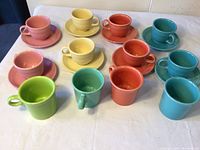 Top-view photo of all cups, saucers, and mugs arranged on a table showing colors and quantity: 2 each of pink, yellow, orange, turquoise cups and saucers; 4 mugs in green, turquoise, orange, and pink.