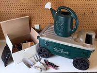 Green wheeled garden cart with beige top, green watering can placed on top, gardening tools and ceramic pots in a white box.