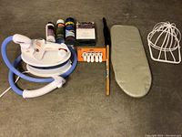 Photo showing Conair clothes steamer, hose, small ironing board, drying rack, plastic clips, brush, and three small bottles of laundry liquids arranged on floor.