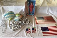 Photo showing patriotic items including boxed Hillary Clinton doll, two paper lanterns, string lights, and two American flags