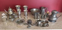 Photo showing an assortment of pewter dishes and serving items arranged on a surface against red wall, highlighting the variety of objects including teapot, goblets, pitchers, candle holders, and smaller accessories.
