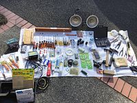 View of full lot laid out on the ground on a white drop cloth. Items include screwdrivers, hammers, brushes, tapes, battery charger, clamps, hardware items, and painting supplies.