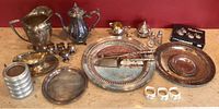 Wide shot of Victorian silver plated serving set including pitchers, platters, small cups, ceramic napkin holders, and boxed salt and pepper set arranged on table.