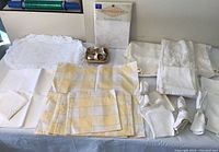 Overview of assorted table linens including folded napkins, placemats with yellow checks, lace overlays, and napkin rings arranged on a white surface.