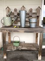 Photo showing the wooden potting table with metal watering can, tall embossed metal vase, two galvanized metal pots, small green planter, gloves, and yellow spigot handles arranged on the table shelves and top.