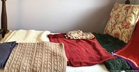 Photo showing multiple woven throw blankets laid out including cream, beige with knit pattern, deep red color. Also visible is a large cushion with floral print.