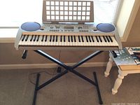Yamaha PSR-275 keyboard on black folding X-stand in front of window, music stand attached on top.