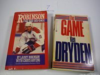 Two vintage hockey books side by side on a white surface: "Robinson for the Defence" with red cover and Larry Robinson hockey player photo, and "The Game" by Ken Dryden with a cream cover and bold text.