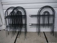 Multiple wire garden fencing stakes with curved tops arranged against white wall on pavement.