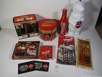Full view of all Coca-Cola collectible items arranged together including tins, figurine, pens, pencils, and cards to show variety in lot.