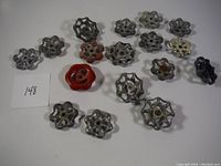 Top-down image showing 16 vintage metal valve handles arranged on a flat surface with a white label '148' visible; handles are mostly silver with one distinctive red handle and one black handle.