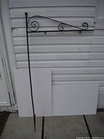 Full view of the wrought iron sign frame against white siding showing the overall shape and size with pointed end for ground installation.