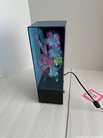 Front view of the fiber optic flower light box showing the colorful artificial flowers inside a rectangular transparent enclosure with a black base and power cord attached.