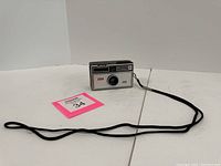 Front view of Kodak Instamatic 100 camera showing lens, viewfinder, Kodak branding, and attached black wrist strap on white background with lot label number 34.