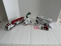 Photo showing five Hess toy vehicles arranged on a white backdrop including firetruck with crane, backhoe, motorcycle, race car, and van.