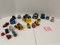 Assorted toy vehicles displayed on white background, showing blue Tonka truck, construction vehicles, Batman car, and other small toys.