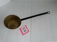 Photo showing side view of old antique copper pan with long straight black metal handle ending in ring.