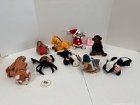 Photo of nine Beanie Babies characters arranged in a white backdrop. Items include a dog, duck, skunk, spider, cow, robin, ape, giraffe, and a cat wearing a Santa outfit.