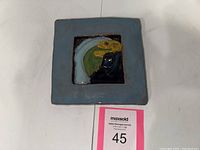 Top-down photo of square ceramic tile with colorful parrot design in relief at center and blue glaze.